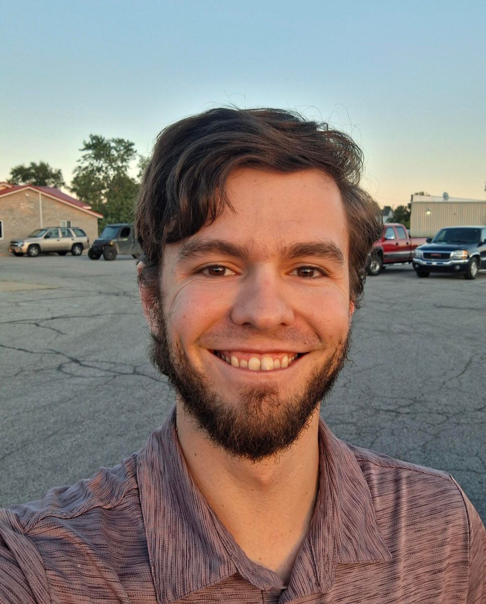 Brendan Johnson smiling in a selfie in a parking lot at dusk, with pickup trucks and a brick building with a red roof behind him